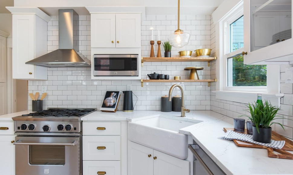 A white kitchen with subway tile, open wood shelving, brass hardware, and a farmhouse sink below a garden window